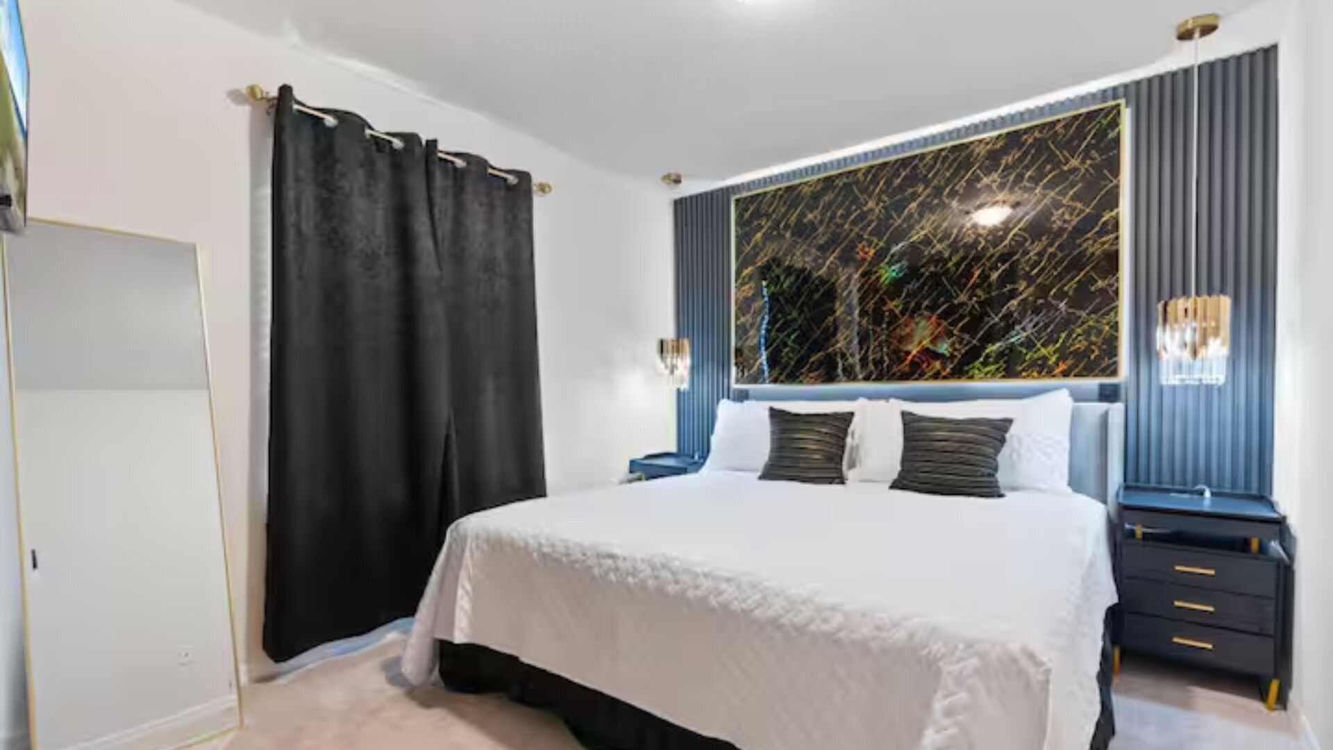 Een moderne slaapkamer met een groot bed, donkere gordijnen, een wandlamp en een decoratieve spiegel.
