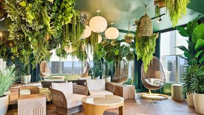 Una sala de estar moderna con techo alto, piso de madera y variedad de plantas. Hay varias zonas de estar, incluyendo un sofá y una silla, además de una mesa de centro redonda.