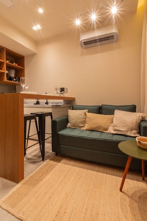 Un espacio habitable compacto con kitchenette, un sofá verde, una mesita redonda y una alfombra tejida.