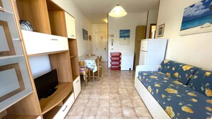 Un espacio habitable compacto con sofá, televisor, estanterías de madera, una mesa de comedor y una zona de cocina.