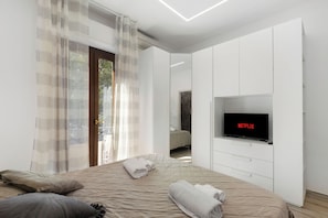 A modern bedroom with a large bed, a flat-screen TV mounted on the wall, and a sliding door leading to a balcony.
