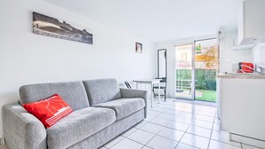 Ein modernes Wohnzimmer mit einem grauen Sofa, einem roten Kissen, einem Essbereich mit Tisch und Stühlen sowie einer Küchenzeile mit einem roten Gerät.