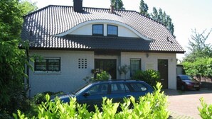 Une maison à deux étages, avec un toit recouvert de tuiles, une voiture garée dans l’allée et, au premier plan, de beaux buissons verts et touffus.