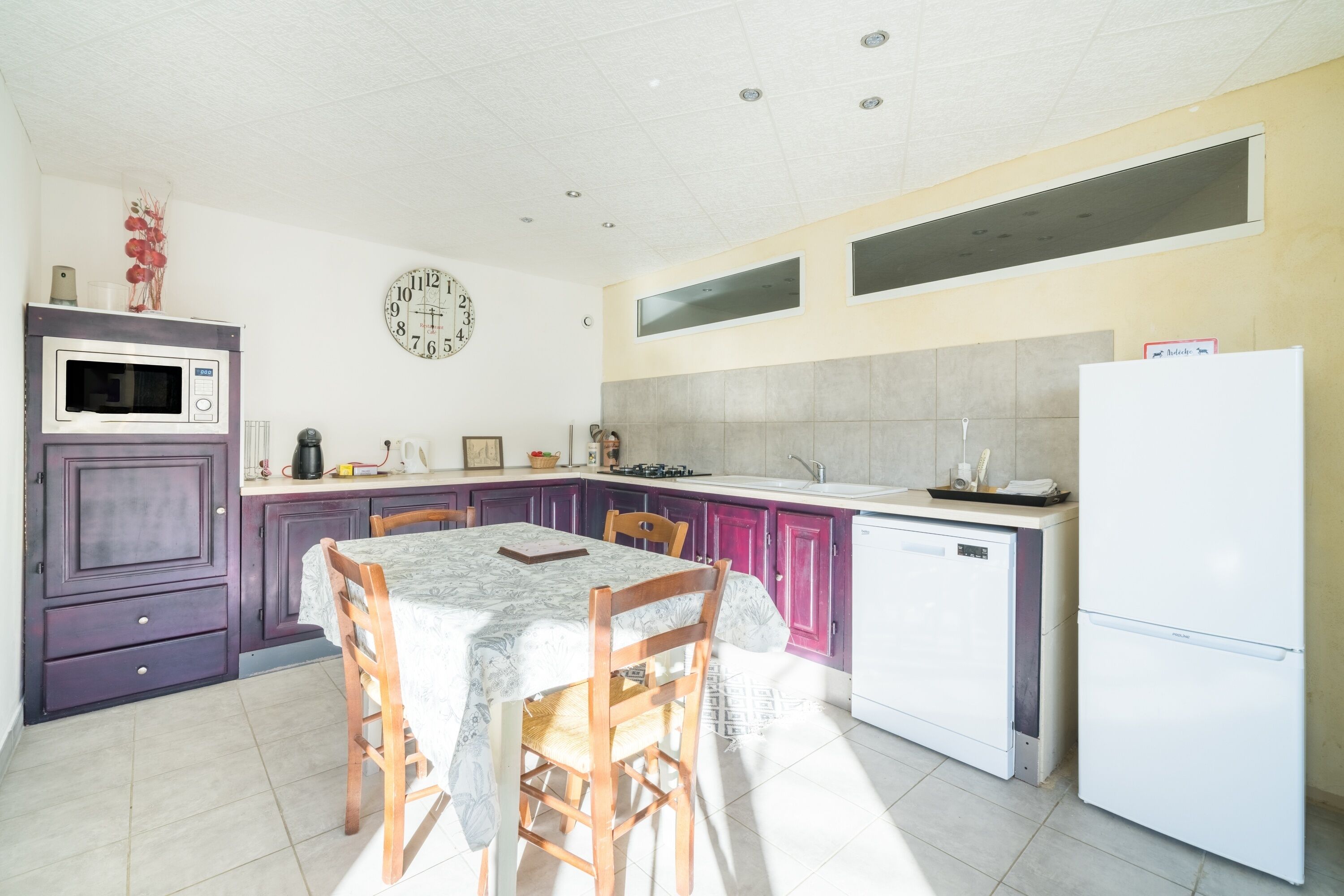 A kitchen with a dining table, chairs, a microwave, a refrigerator, and a clock on the wall.
