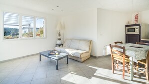 Ein Wohnzimmer mit einer Couch, einem Couchtisch und einem Esstisch mit Stühlen. Ein Fenster mit Jalousien, eine Lampe und eine Mikrowelle sind an der Wand angebracht.