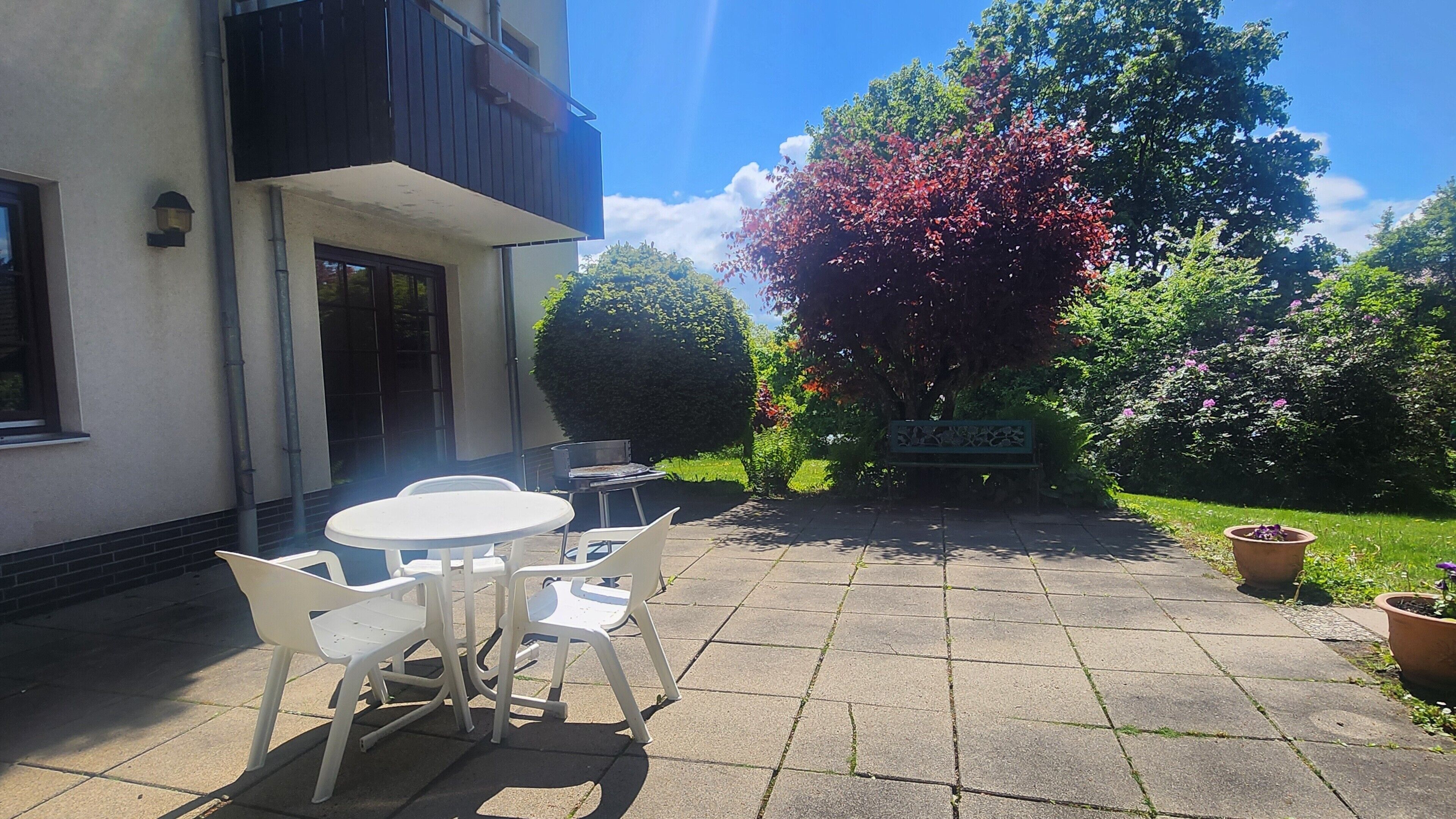 An outdoor patio with a round table and chairs, a bench, and a garden with trees and shrubs.