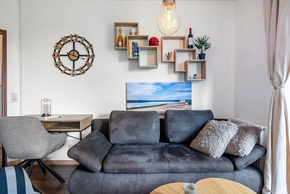 A modern living room with a grey sofa, a wooden coffee table, a desk with a lamp, and a wall clock.