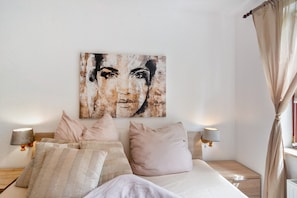 A bedroom with a bed, pillows, a wall-mounted artwork, bedside lamps, and a window with curtains.