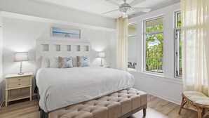 Quarto com cama, mesas de cabeceira, ventoinha no teto, janela com cortinas e vista para o verde lá fora.