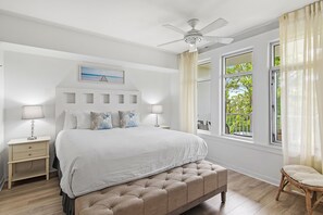 Ein Schlafzimmer mit Bett, Nachttischen, Deckenventilator, Fenster mit Vorhängen und Blick auf Grünfläche.