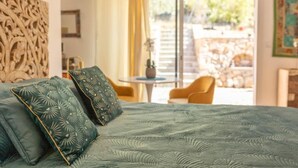 Una cama con una colcha y almohadas estampadas, un cabecero de madera, vista a un patio con sillas y una mesa, y una puerta que da al exterior.