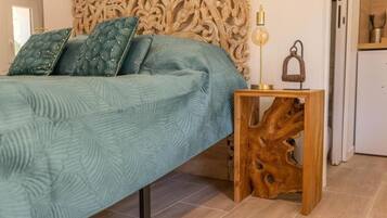 A bed with a textured teal bedspread, a carved wooden headboard, a small wooden side table with a lamp, and a rustic wooden base.