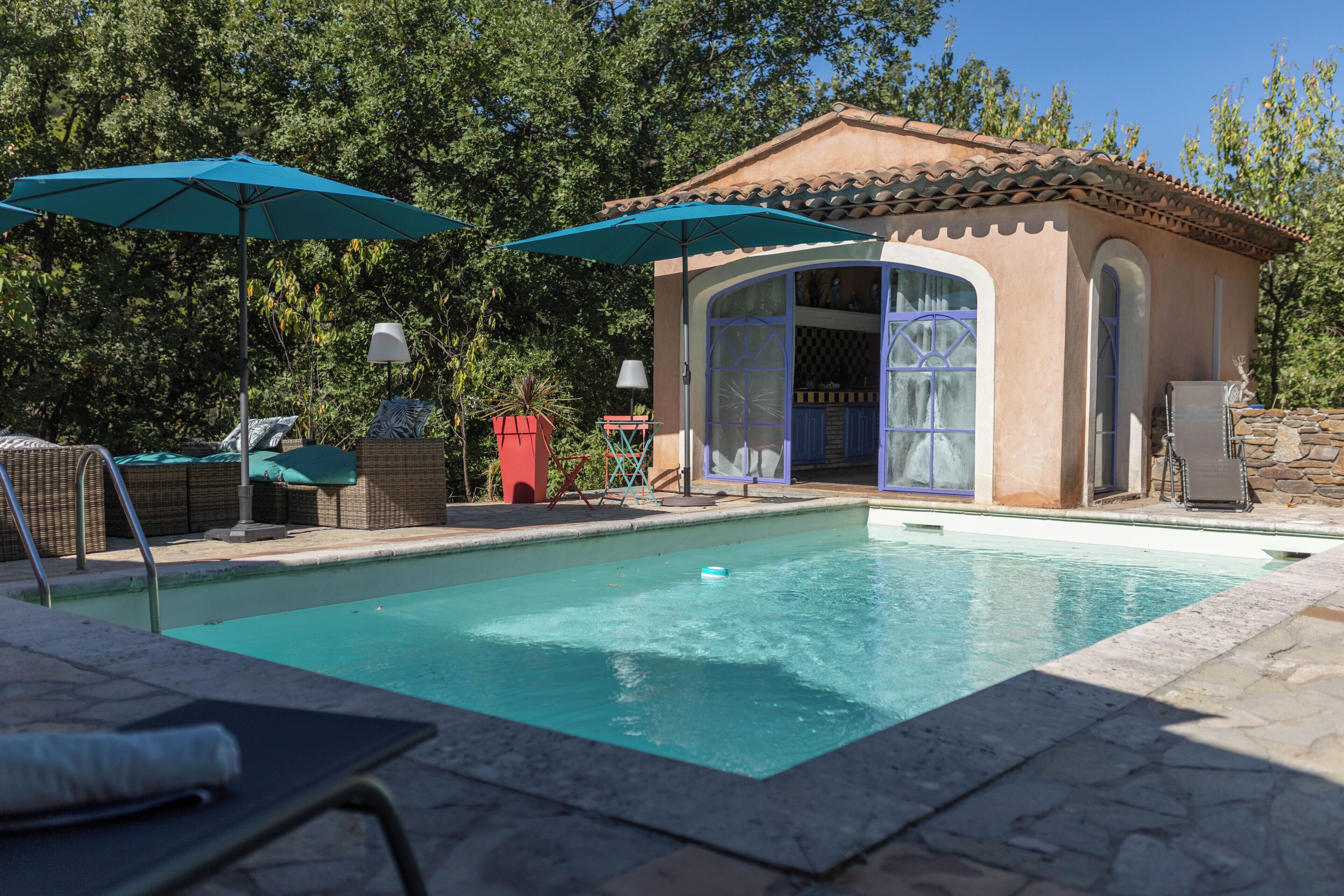 Une piscine avec des chaises longues et des parasols, un petit bâtiment doté d’un toit en tuiles et un mur en pierre.