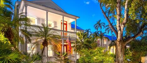 A two-story colonial-style building with a balcony, surrounded by lush tropical vegetation and a clear blue sky.
