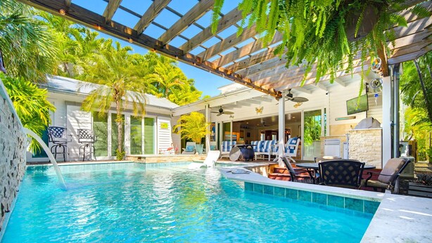 Ein Swimmingpool mit Liegestühlen und Sonnenschirmen, umgeben von Palmen und einer Pergola.