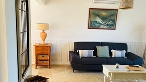 Una sala de estar con un sofá oscuro, una mesa de centro blanca, una mesita de noche de madera con una lámpara y un cuadro de una ola en la pared.