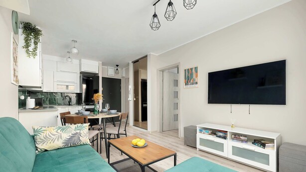 A modern living room with a sofa, a coffee table, and a TV. There is a kitchen area with cabinets and a dining table. - Comfortable Apartment | Garden and Private Parking (Szczecin)