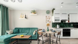 A modern living room with a teal sofa, a wooden coffee table, and a dining area with chairs and a table. A kitchen with white cabinets and a green backsplash is visible in the background. - Comfortable Apartment | Garden and Private Parking (Szczecin)