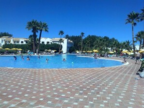 Una piscina con suelo de baldosas, palmeras y sombrillas.