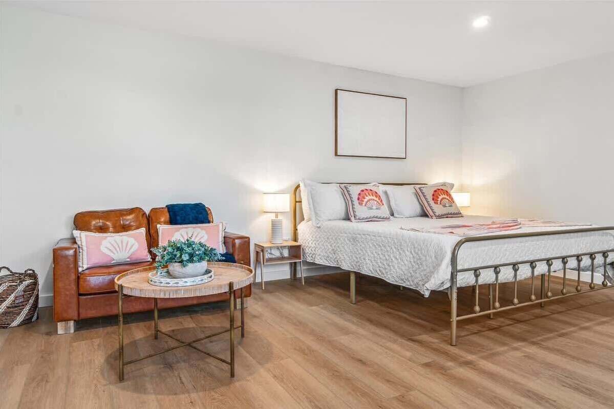 Un dormitorio con una cama, un sofá de cuero marrón, una pequeña mesa redonda con una planta y una mesita de noche con una lámpara.