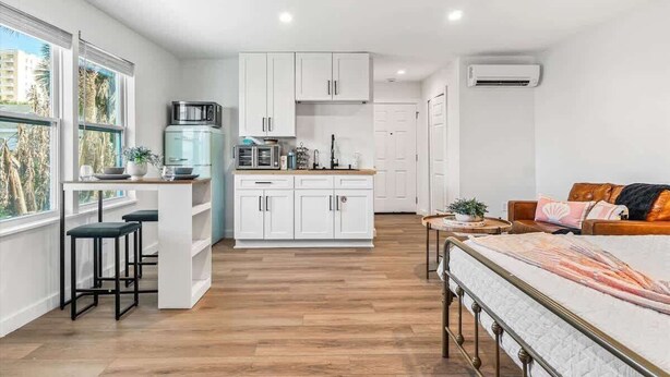 A compact studio apartment with a kitchenette, a dining area, a bed, and a large window providing a view of palm trees.