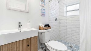 Et moderne badeværelse med hvid vask, toilet og brusebad. To billeder hænger på væggen, og der er en kurv med et håndklæde på toilettet.