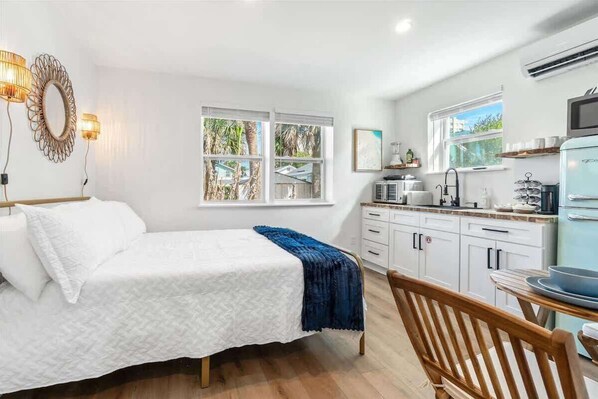 A bedroom with a bed, a wooden chair, a desk with a chair, a kitchen area with a refrigerator, and a window with a view of palm trees.