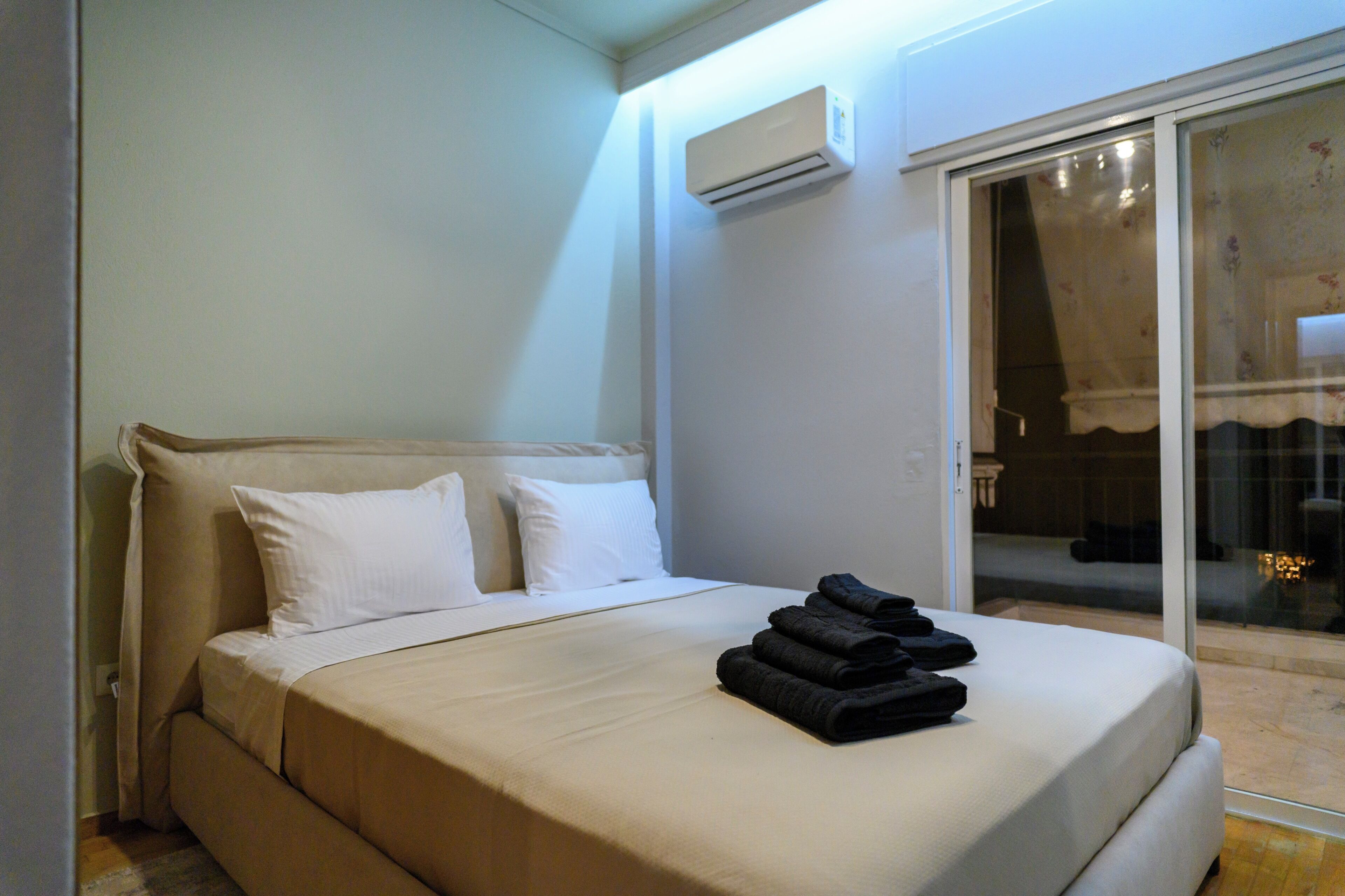 A hotel room with a bed, white pillows, a folded towel, an air conditioning unit, and a sliding glass door leading to an outdoor area.