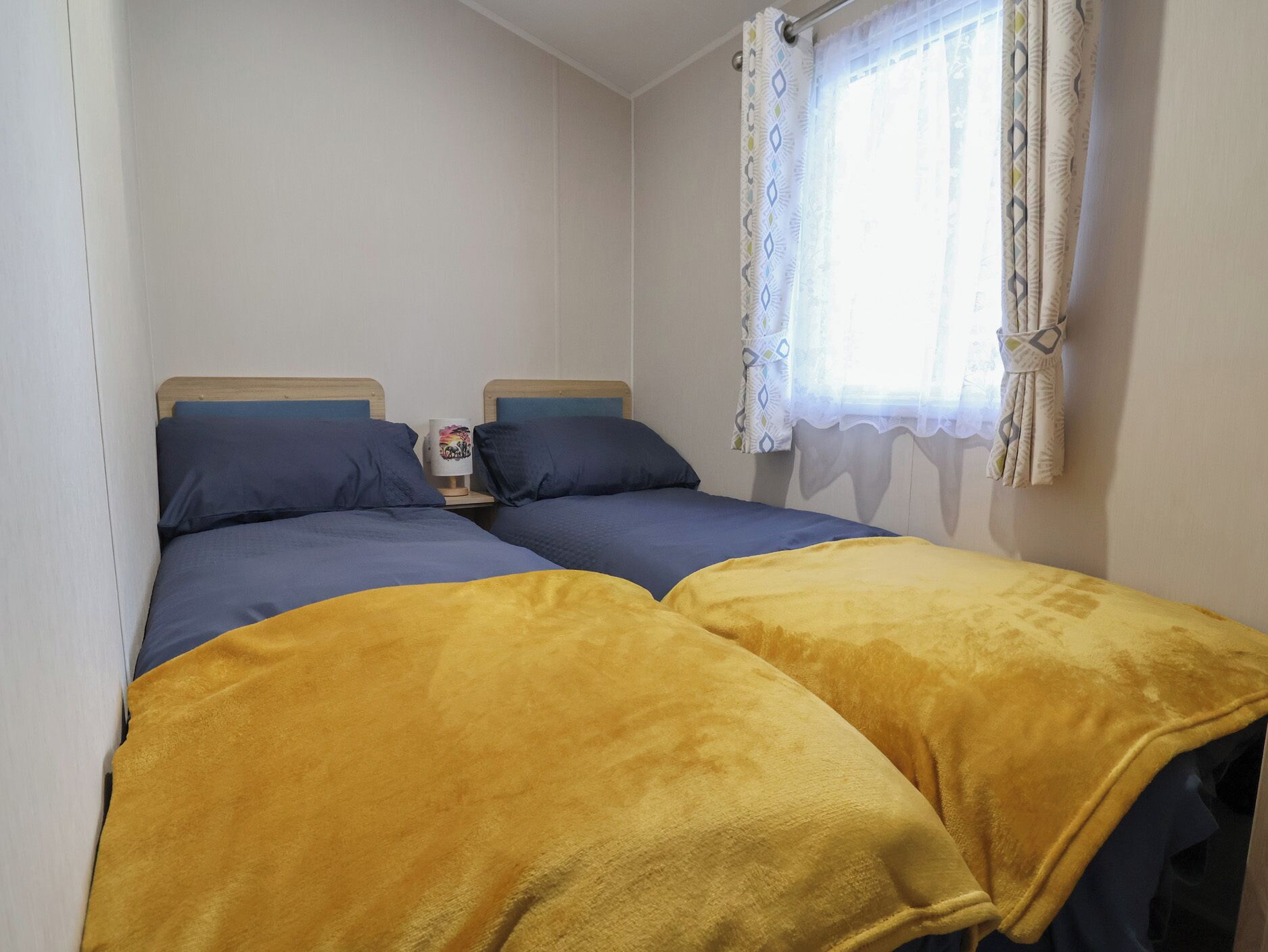 Un dormitorio con dos camas, una ventana con cortinas estampadas y una mesita de noche con una lámpara.