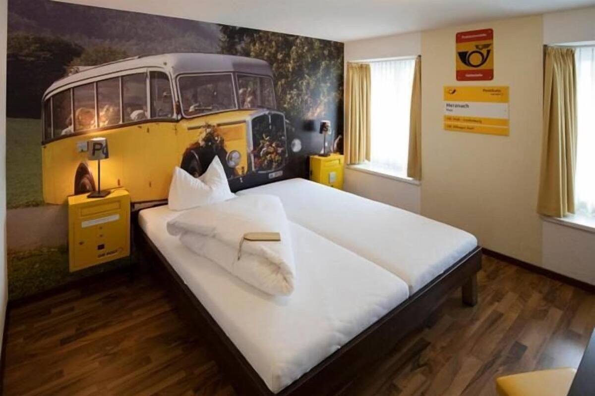 A hotel room with a bed, a nightstand, a wall-mounted lamp, a window with curtains, and a wall decoration of a vintage bus.