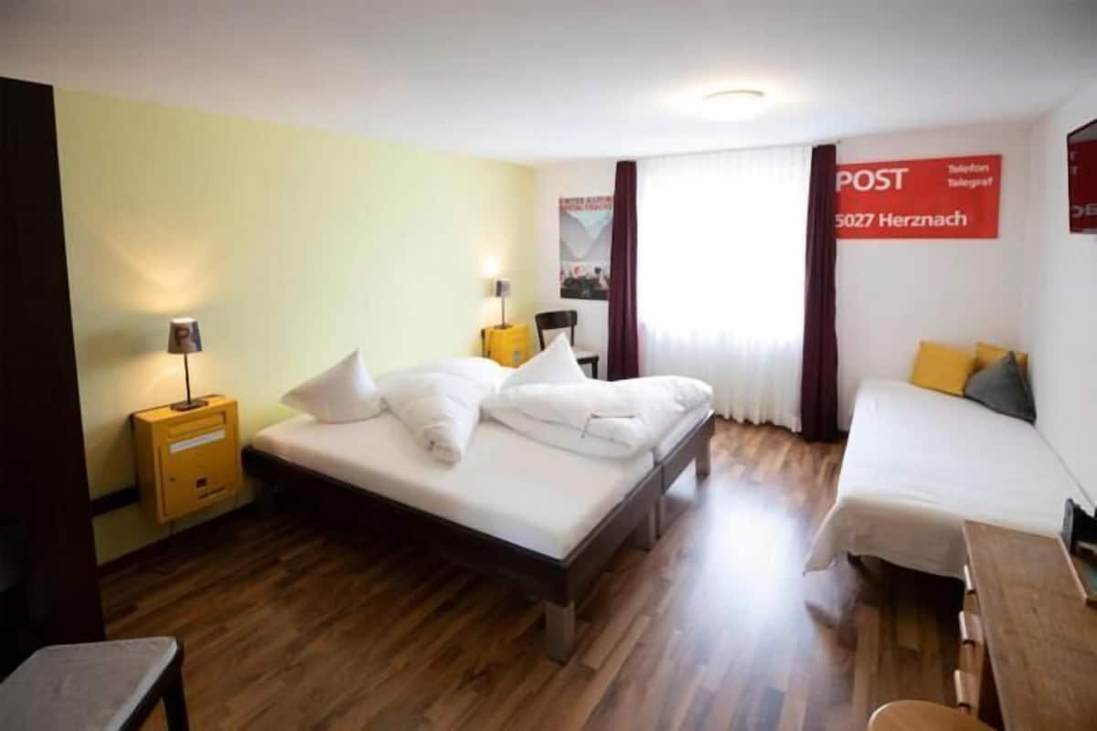 A hotel room with a bed, a bedside table, a chair, a desk, and a window with curtains.