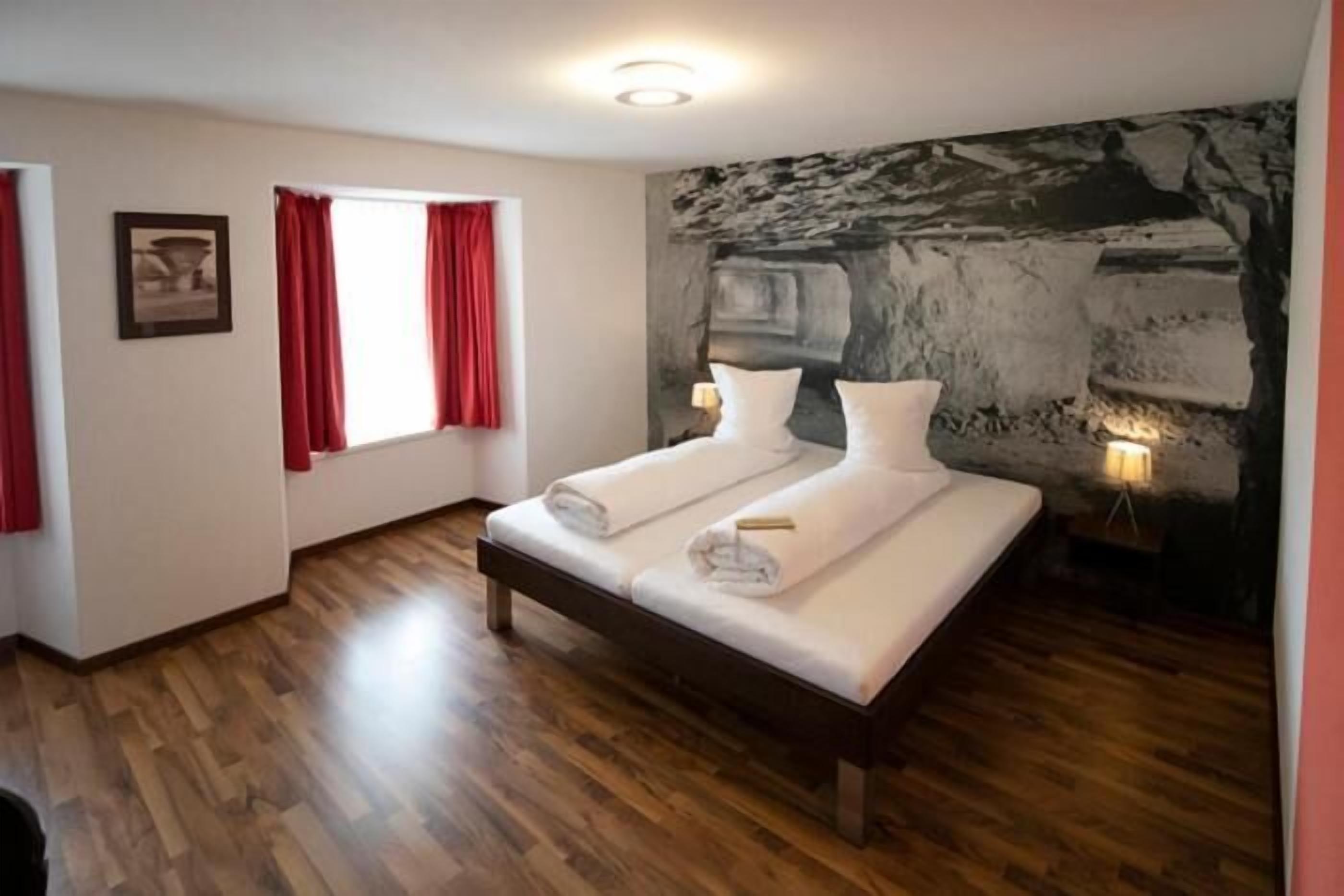 A bedroom with a bed, a window with red curtains, a stone wall, and a wooden floor.