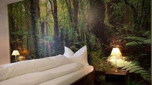 Un letto con biancheria da letto bianca in una stanza con un murales a tema foresta, due abat-jour e un comodino in legno.
