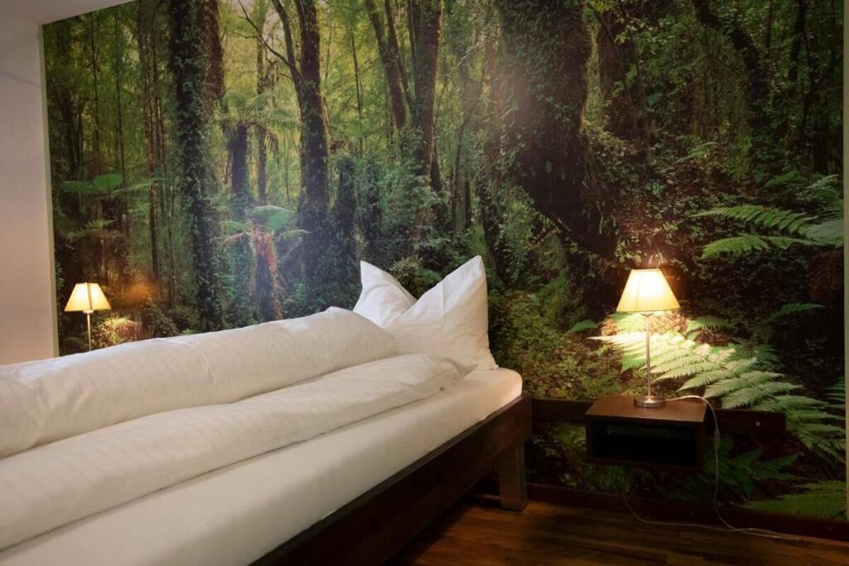 A bed with white bedding in a room with a forest-themed wall mural, two bedside lamps, and a wooden nightstand.