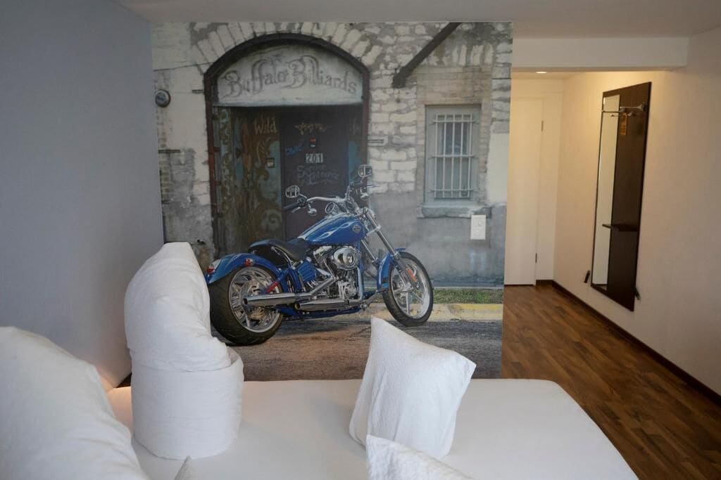 Un dormitorio con un mural de una motocicleta, una cama con ropa de cama blanca y suelo de madera.