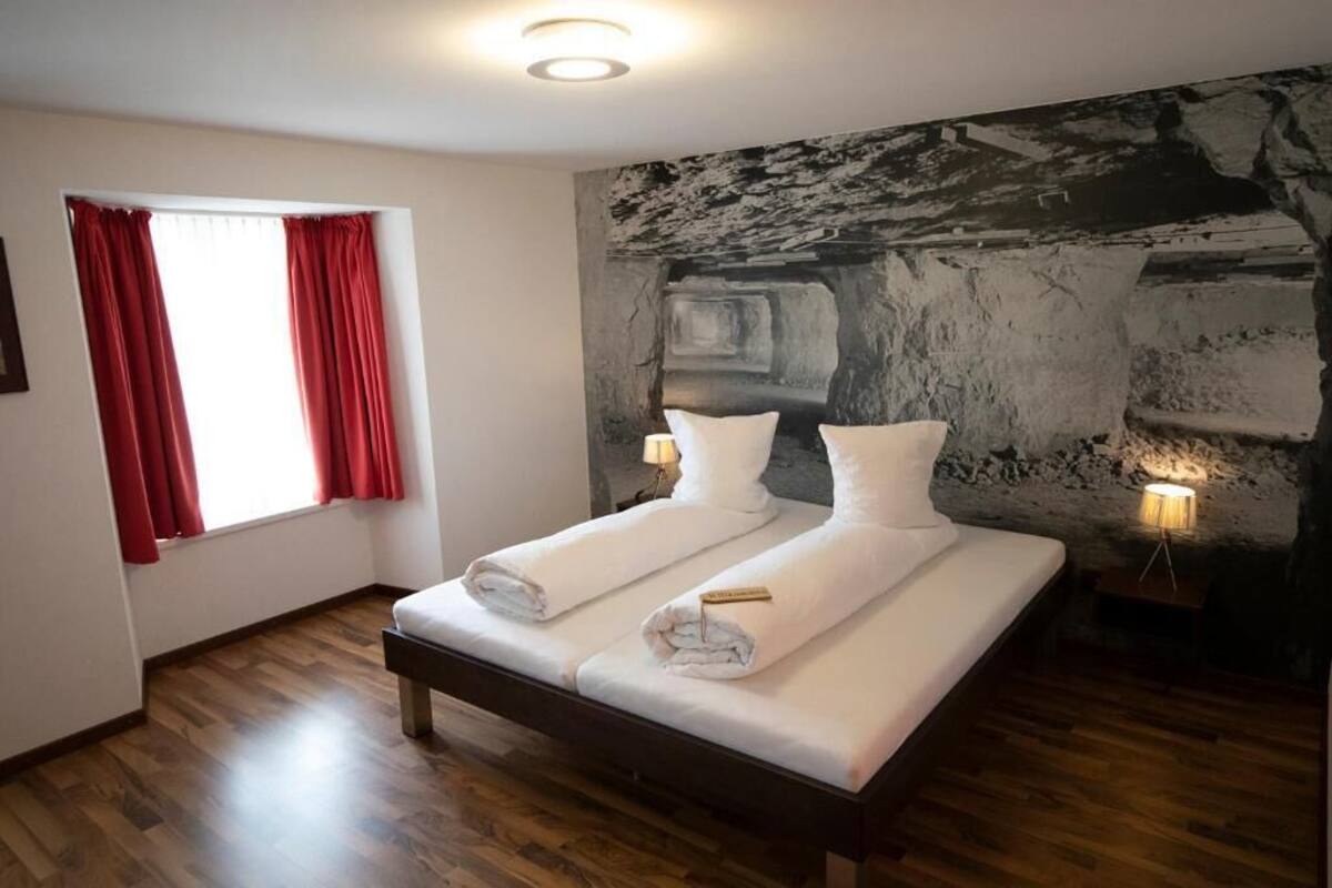 A room with a bed, two bedside lamps, a window with red curtains, and a textured wall.