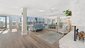 A spacious living room with a blue sofa, a white fireplace, and large windows offering a view of the outdoors. - Gulffront Condo w/ Tiki Bar, Pools, & Sunsets (Panama City Beach)