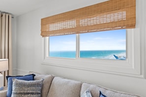 Une pièce dotée d’une grande fenêtre donnant sur l’océan, d’un canapé beige agrémenté de coussins à motifs et d’une lampe blanche posée sur une table d’appoint.