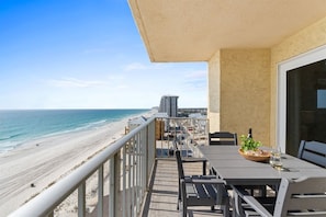 A balcony with a view of the beach and ocean, a table with chairs, and a bottle of wine. - Gulffront Condo w/ Tiki Bar, Pools, & Sunsets (Panama City Beach)