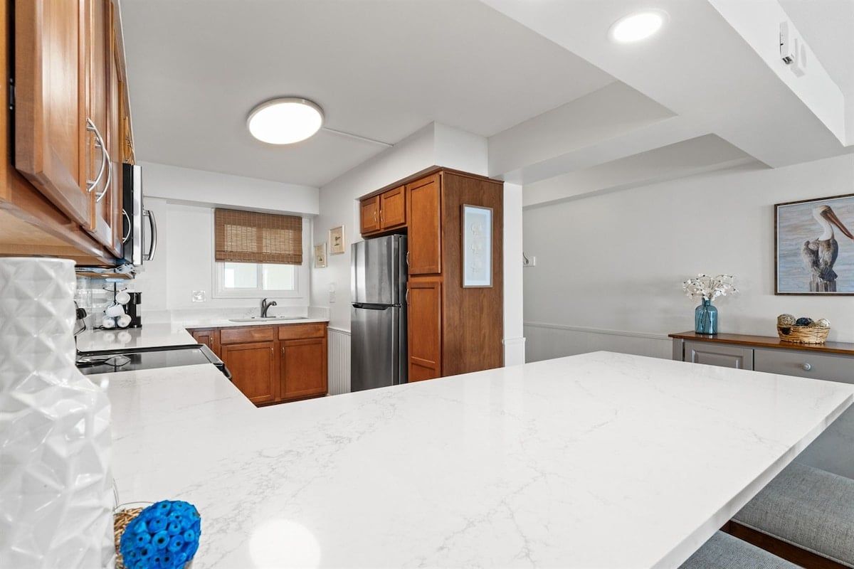 A modern kitchen with white marble countertops, wooden cabinets, a stainless steel refrigerator, and a large island.