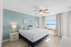 A bedroom with a bed, bedside tables, a ceiling fan, a window with curtains, and a view of the sea. - Gulffront Condo w/ Tiki Bar, Pools, & Sunsets (Panama City Beach)