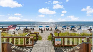 A wooden deck leading to a beach with umbrellas and lounge chairs, an American flag, and caution signs. - Gulffront Condo w/ Tiki Bar, Pools, & Sunsets (Panama City Beach)