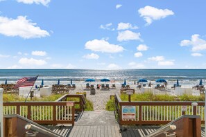 Une terrasse en bois menant à une plage agrémentée de parasols et de chaises longues, avec un drapeau américain et des panneaux d’avertissement.