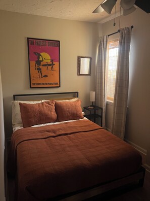 Een slaapkamer met een bed, een poster aan de muur, een nachtkastje en een raam met gordijnen.
