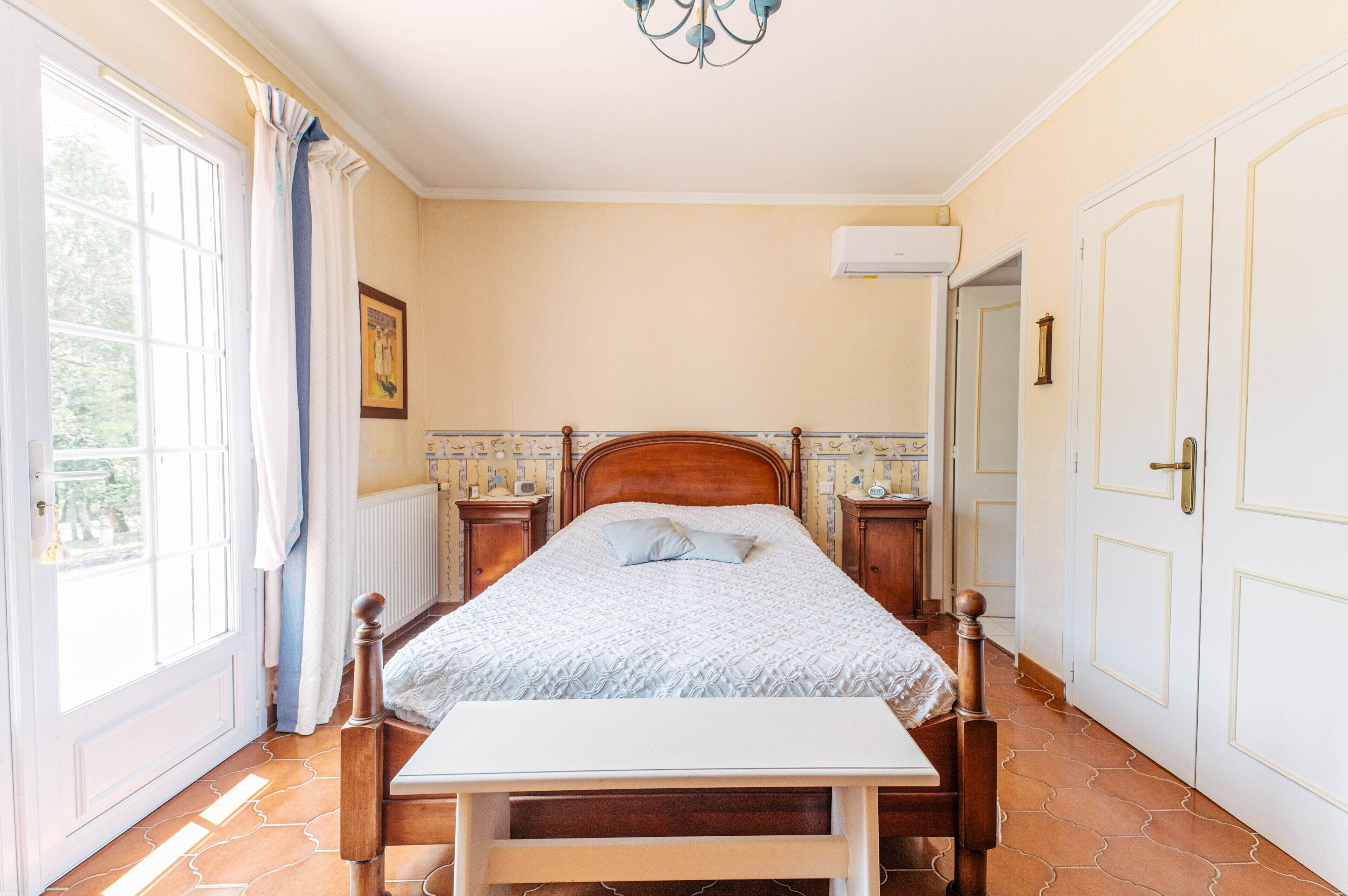 A bedroom with a wooden bed, a bedside table, a window with curtains, a radiator, and a door.