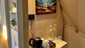 Ein kompaktes Hotelzimmer mit Bett, Nachttischlampe, einem kleinen Tisch mit Wasserkocher, Gläsern und Flaschen, sowie einem Gemälde einer Berglandschaft an der Wand.