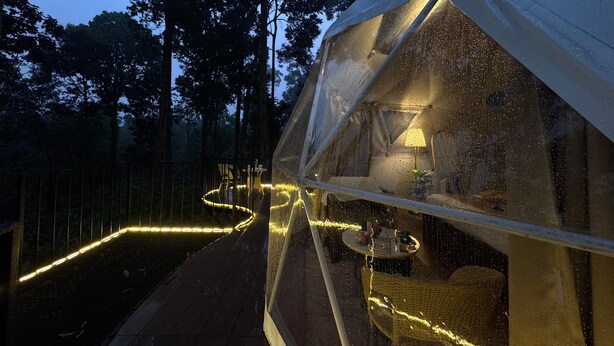 Ein durchsichtiges Zelt mit Tisch und Stuhl im Inneren, umgeben von einer Holzterrasse und beleuchtet mit Lichterketten.