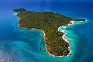 An aerial view of a tropical island with clear turquoise waters, dense green vegetation, and a narrow sandy beach.