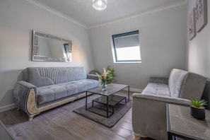 A modern living room with a grey sofa, a glass coffee table, and a grey armchair. A large mirror hangs on the wall, and there is a window with a blue frame. - Modern Flat in Waterloo - Central London (London)