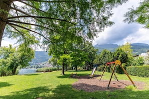 A park with a playground, including swings and a slide, surrounded by trees and a lake view. - Mobile Home at Lake Caldonazzo (Pergine Valsugana)